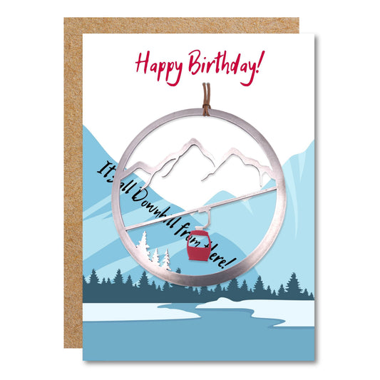 Birthday ornament card shows a a mountain range with the inscription happy birthday, it&#39;s all downhill Fromm there!. Attached is an ornament of a mountain gondola